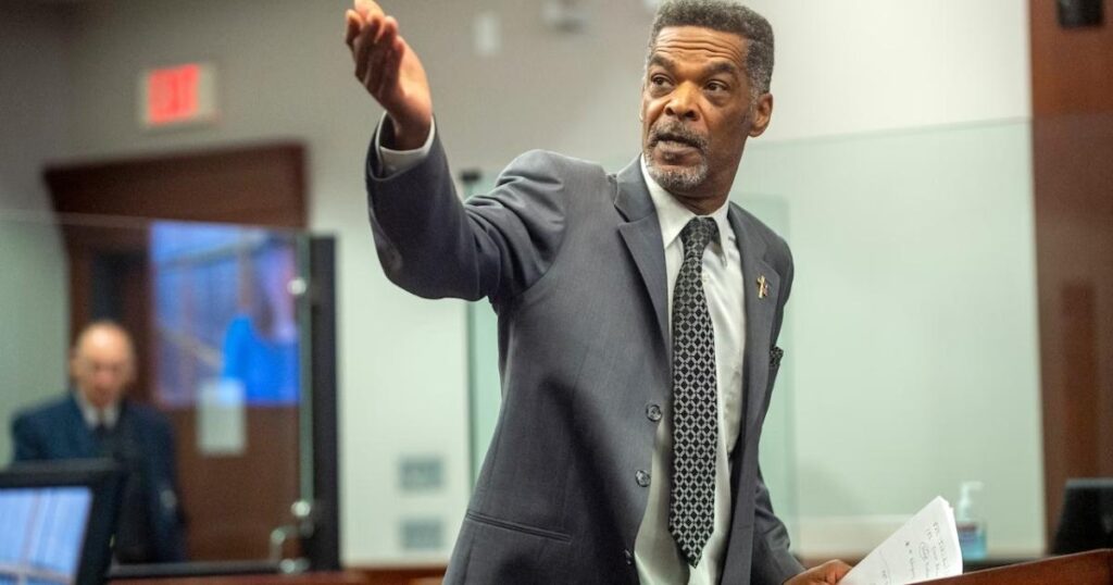 Man in a suit gesturing while speaking in a courtroom setting.