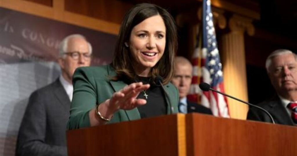 A person gestures while speaking at a podium with others in the background, in a conference room.