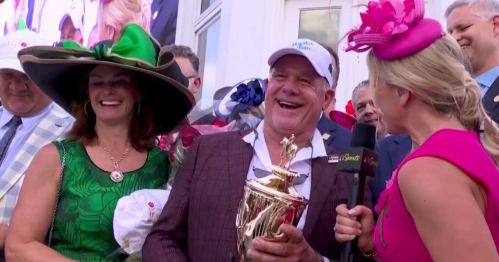 Group of elegantly dressed people at an event with a trophy, one woman holding a microphone.