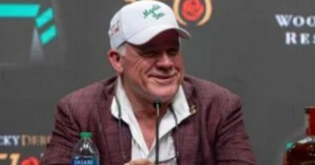 Person in a cap and suit sitting at a press event with microphones and bottled water.