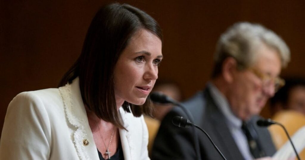 A woman in a white jacket sitting in a hearing room with microphones, in front of blurred individuals.